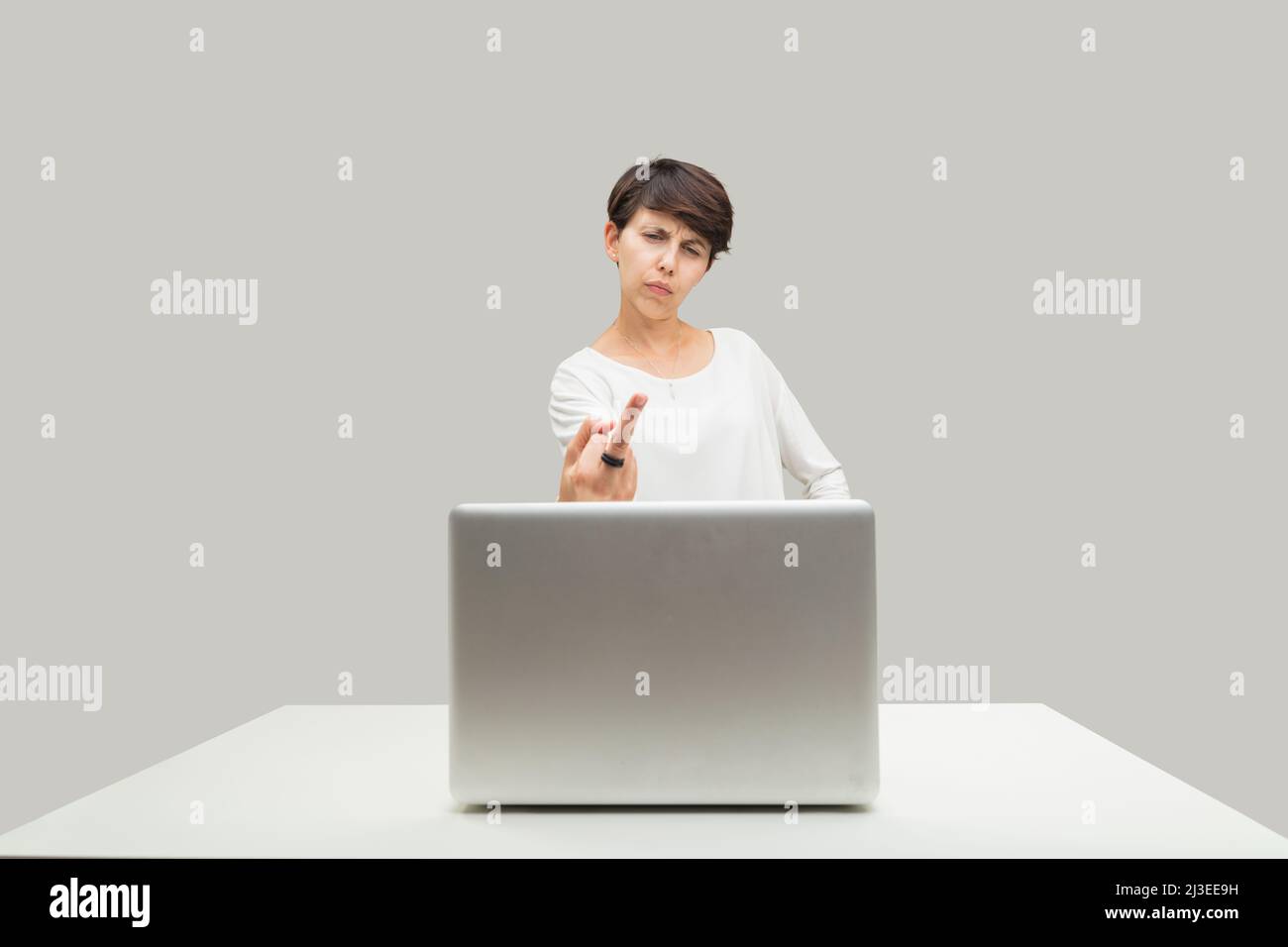 Angry ordinary young woman gestures toward her laptop screen, erects middle finger toward what she disagrees with: injustice, evil Stock Photo