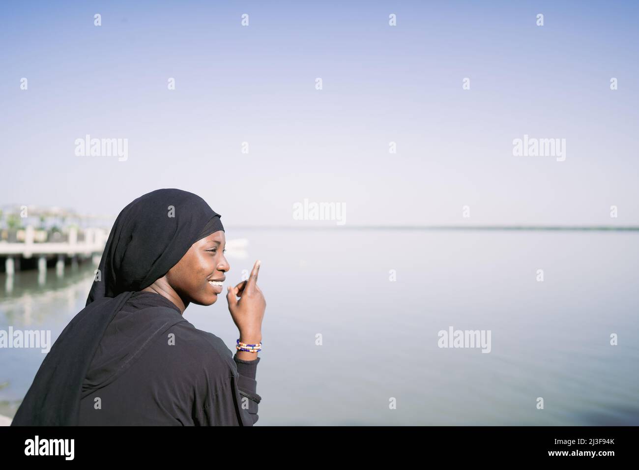 Smiling African Muslim girl sitting relaxed by the sea pointing to the horizon, symbolizing trust and hope in the future of young generations Stock Photo