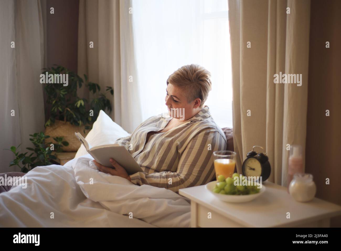 Happy overweight woman reading book in bed at home. Stock Photo