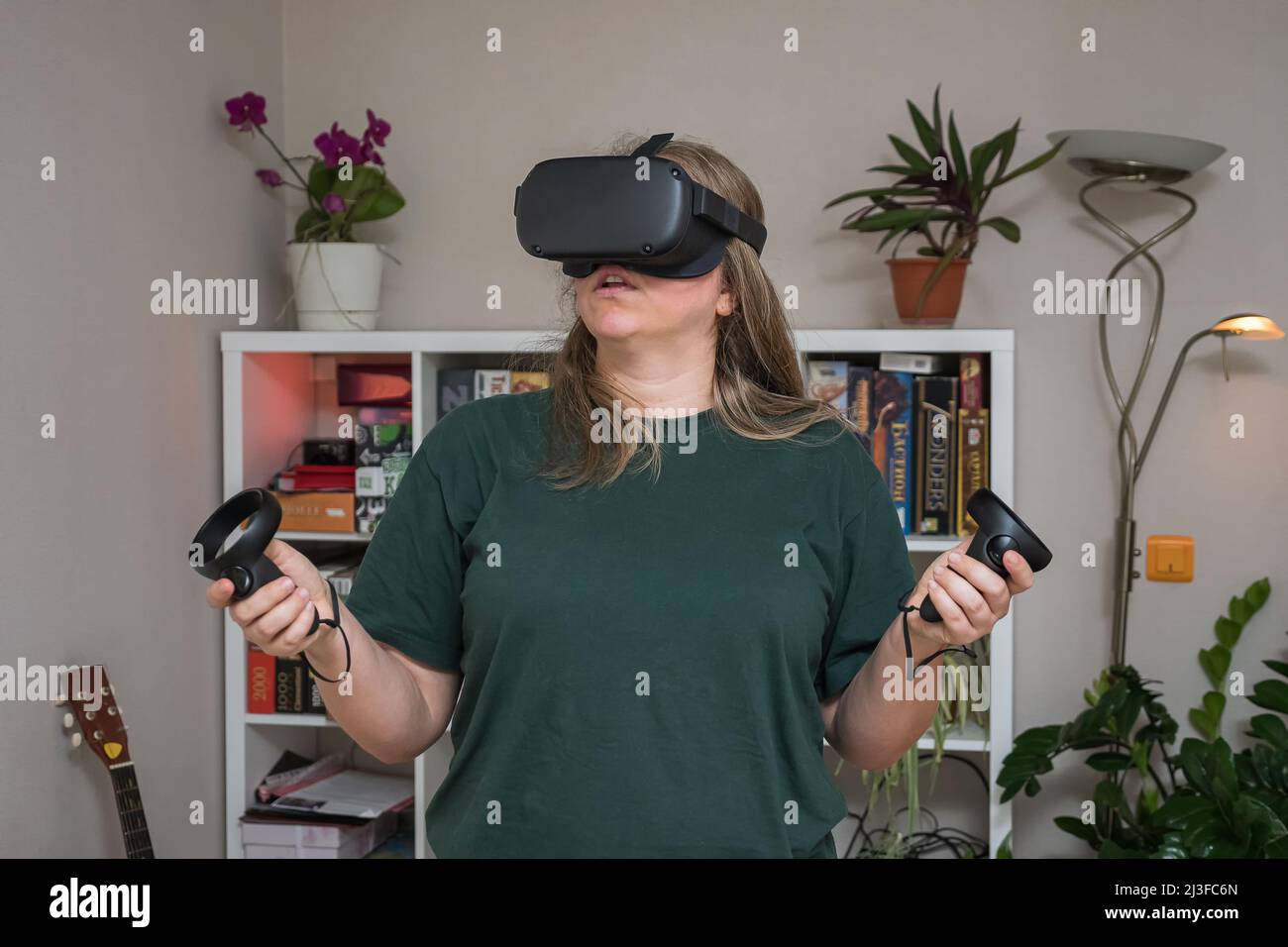 A girl in virtual reality glasses, with controllers in her hands in a gray room in front of a rack of board games. VR helmet for gaming, education. Kr Stock Photo