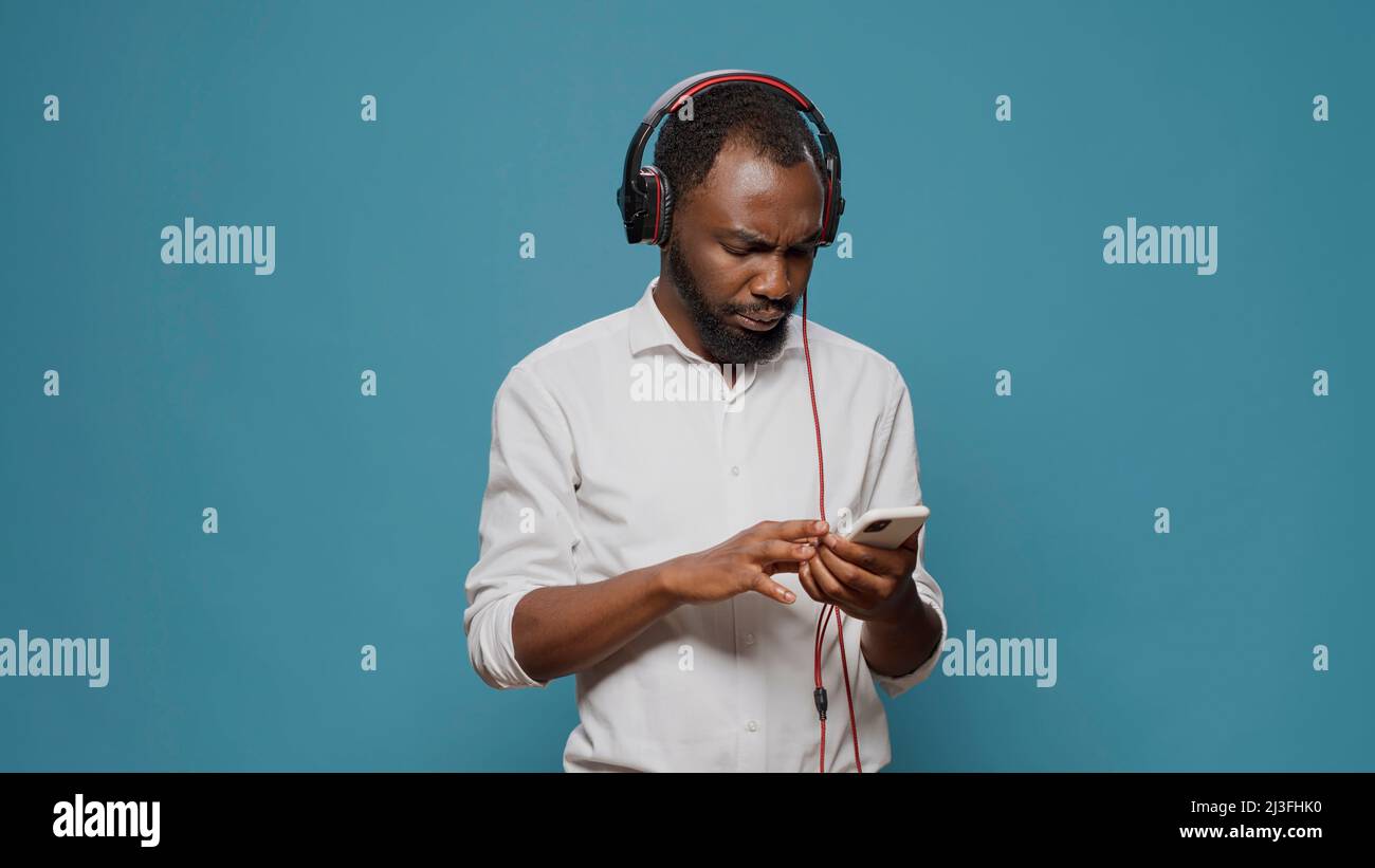 Young person listening to sound on headphones with mobile phone, paying attention to podcast. Confused man feeling uncertain about internet information on smartphone and audio heaset. Stock Photo