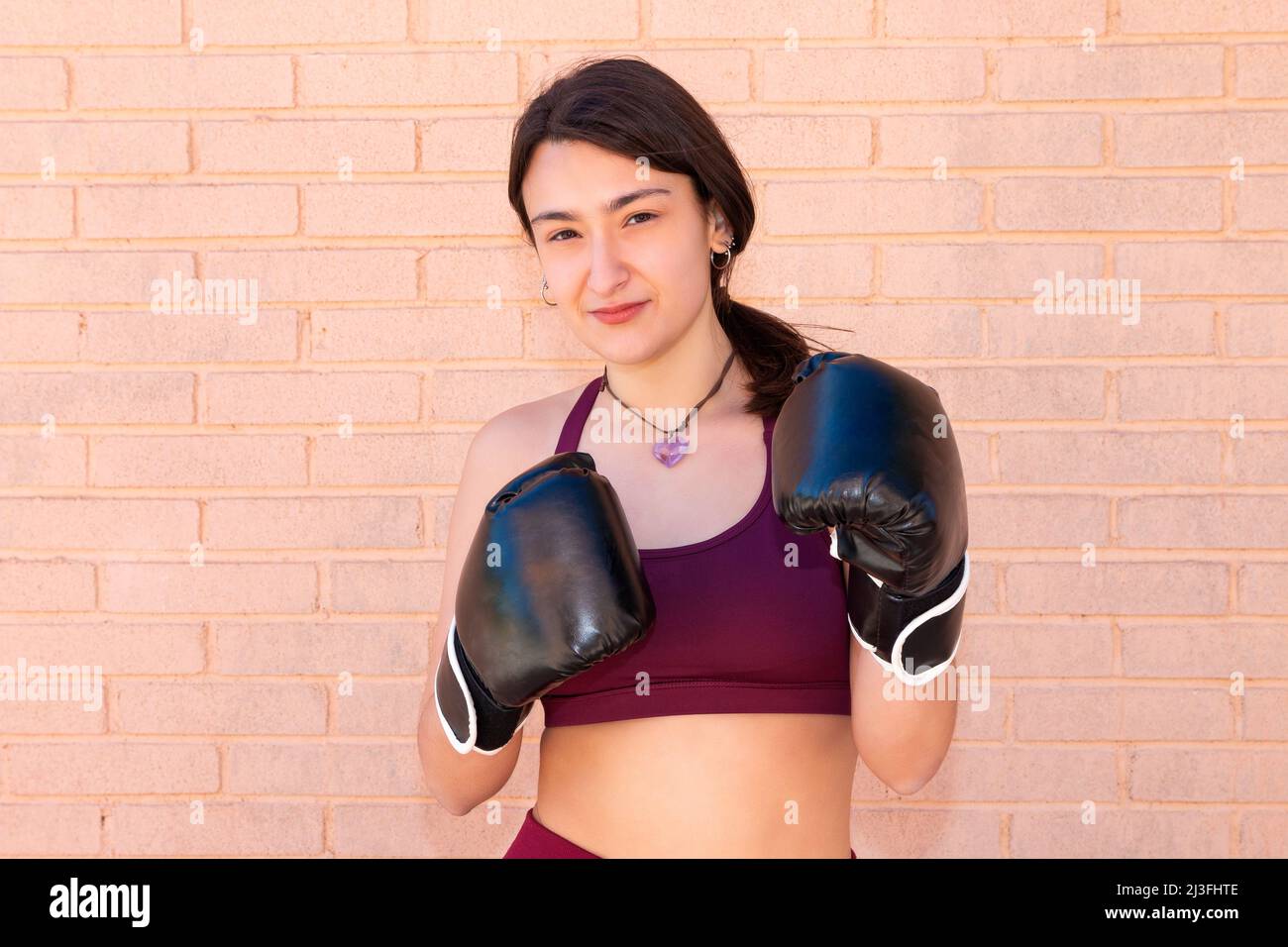 A young Caucasian woman wearing black boxing gloves has her arms flexed with her fists close to her chest. in the background is a brick wall. Stock Photo