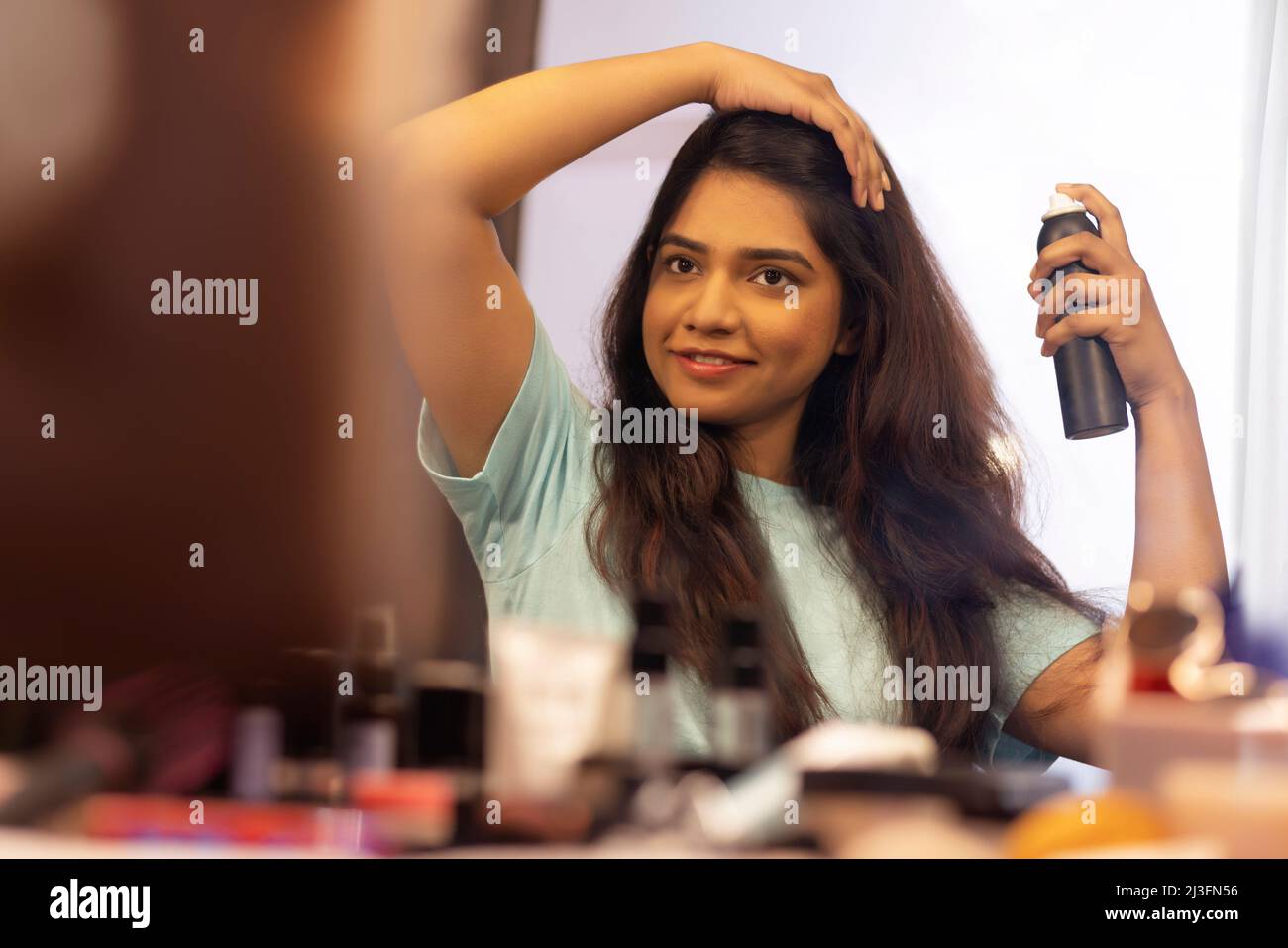 Young woman applying hair spray on her hair in front of mirror Stock Photo