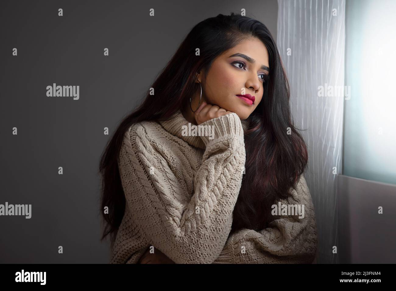 Young woman in winter cloth sitting near window and starring out with hand on chin Stock Photo