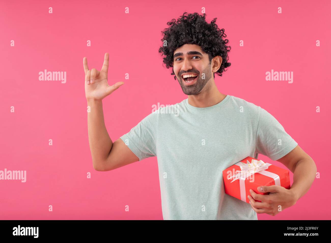 Funny young man with false wig looking at camera with gift and gesturing with fingers Stock Photo