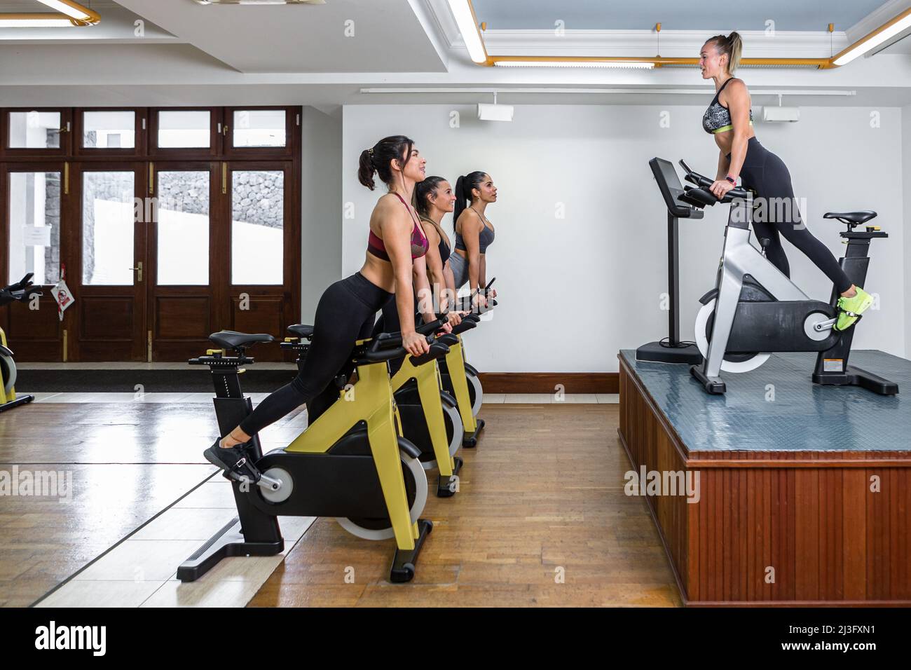 Side view of sportswomen and female instructor leaning on handlebar of exercise bike during fitness training in light gym Stock Photo