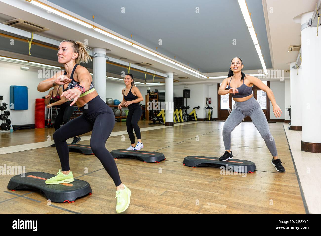 Group of fit female athletes in activewear smiling and lunging on steppers during aerobic workout in spacious gym Stock Photo