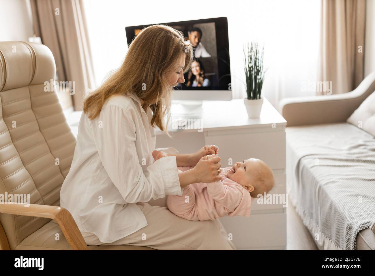 Young woman working at desk at home with computer holding crying tired infant baby holding business meeting at home, online pediatric consultation, ch Stock Photo