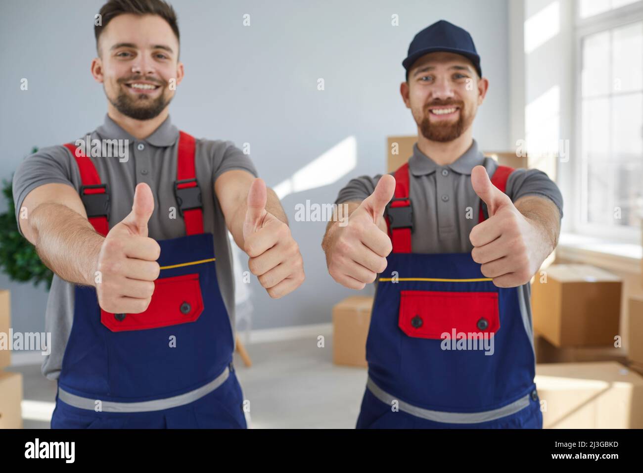 Two smiling young male employees of moving and cargo delivery company show thumbs up. Stock Photo
