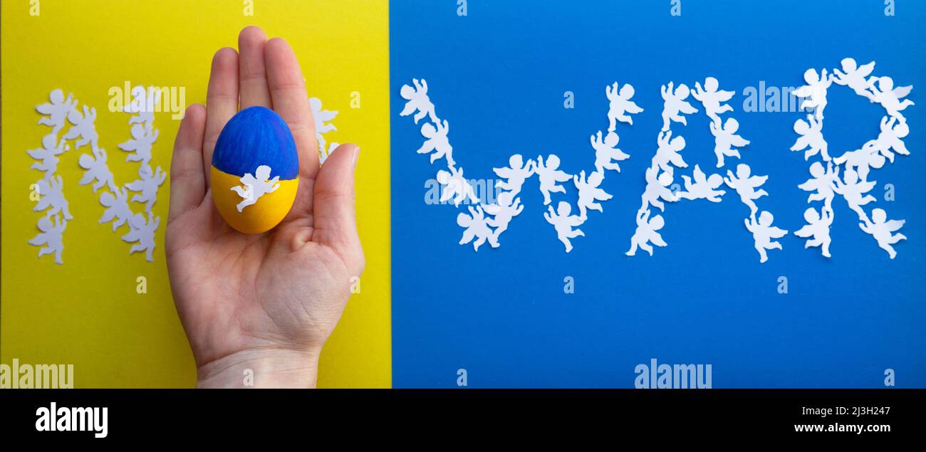 inscription - no war, lined with paper angels on a yellow-blue background. Easter egg painted in colors of Ukrainian flag in hand instead of letter o. Stock Photo