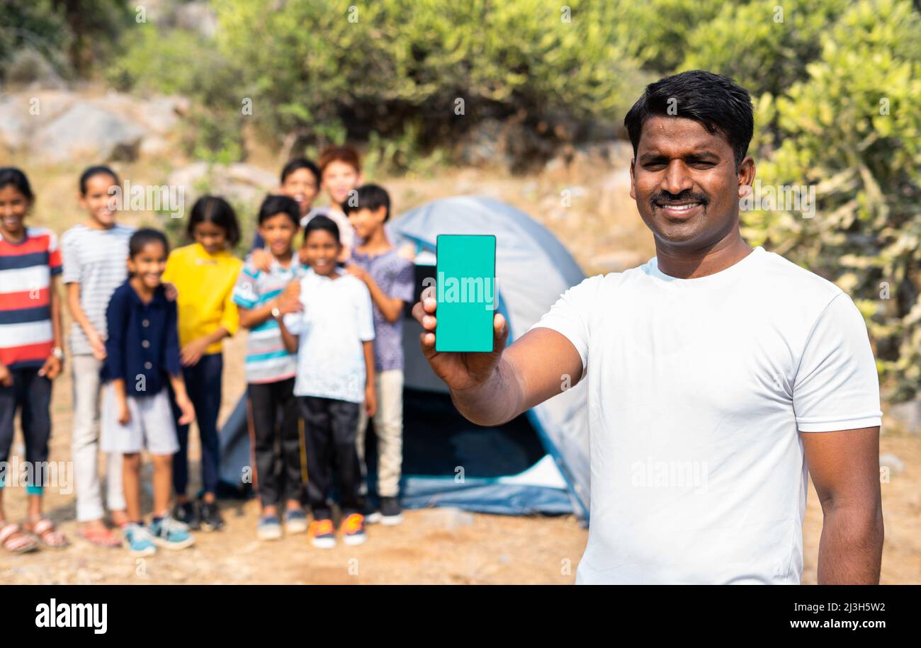 Teacher showing green screen mobile phone by looking camera at summer camp while kids playing background in front of tent - concept of app promotion Stock Photo