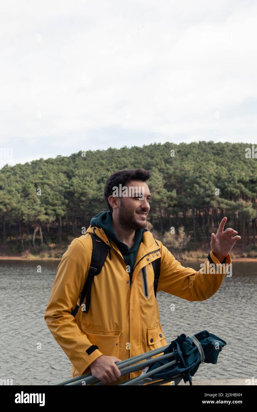 A boy with yellow raincoat standing in front of the lake. He has camp chair on his hand. He will have a rest by the lake. Nature park on a gold autumn Stock Photo