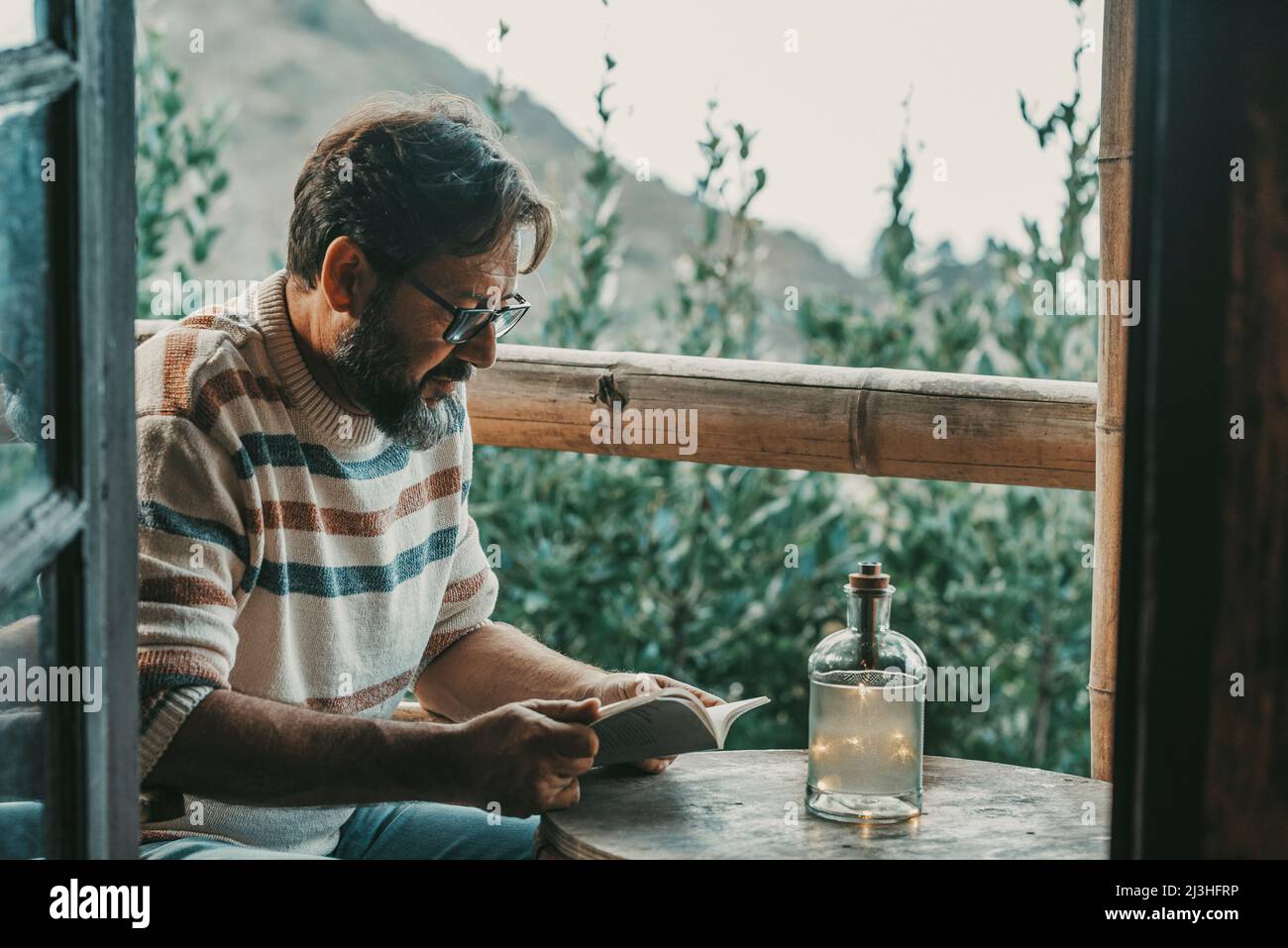 Mature adult man sitting at home in the outdoor terrace reading a book and relaxing enjoying the nature outside in background - relaxed male people enjoy read and study - bearded portrait of man Stock Photo