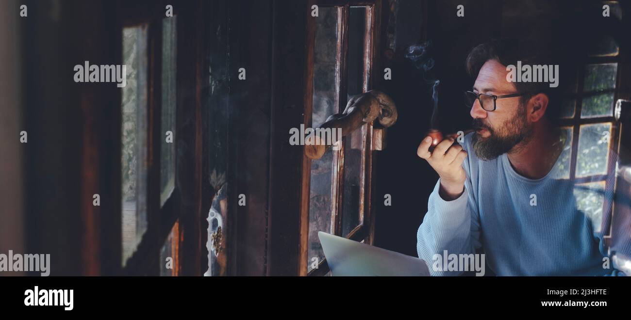 Mature man at work on laptop computer sitting at the table inside a home cozy cabin with wooden windows - digital smart working adult people concept lifestyle - smiging pipe Stock Photo