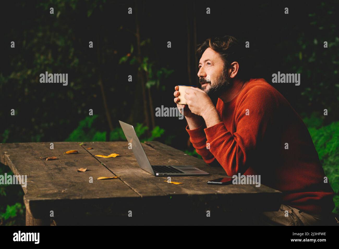 Concept of remote worker and smart working digital modern people lifestyle with adult hipster bearded man work outdoors on a table in the woods and drinking coffee thinking about job - online internet connection laptop computer Stock Photo