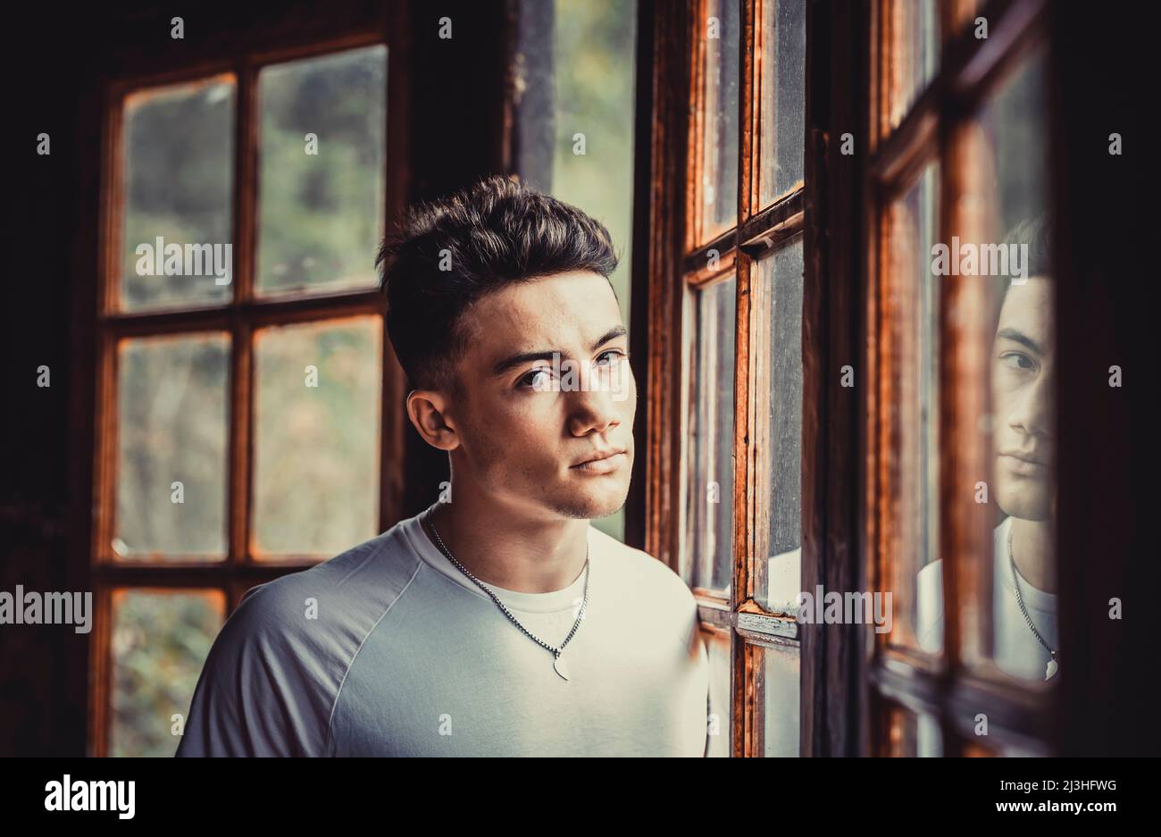 Portrait of serious boy teenager age looking on camera near cozy chalet wooden windows - concept of teen problems life indoor Stock Photo