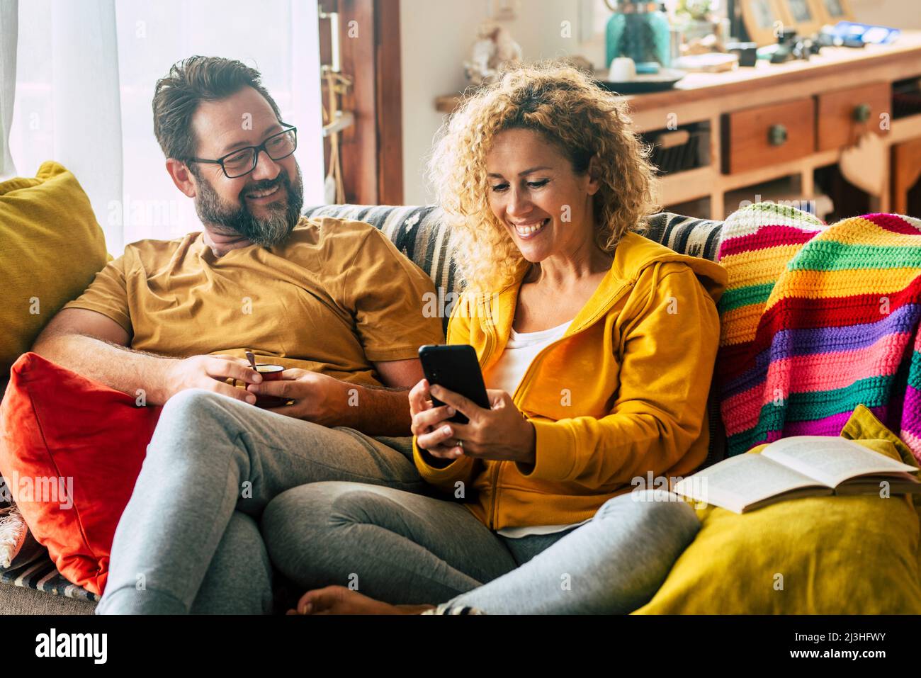 Adult couple at home enjoy mobile phone activity sitting on the sofa and smiling. Colorful house interior and cheerful man and woman people having morning breakfast with technology Stock Photo