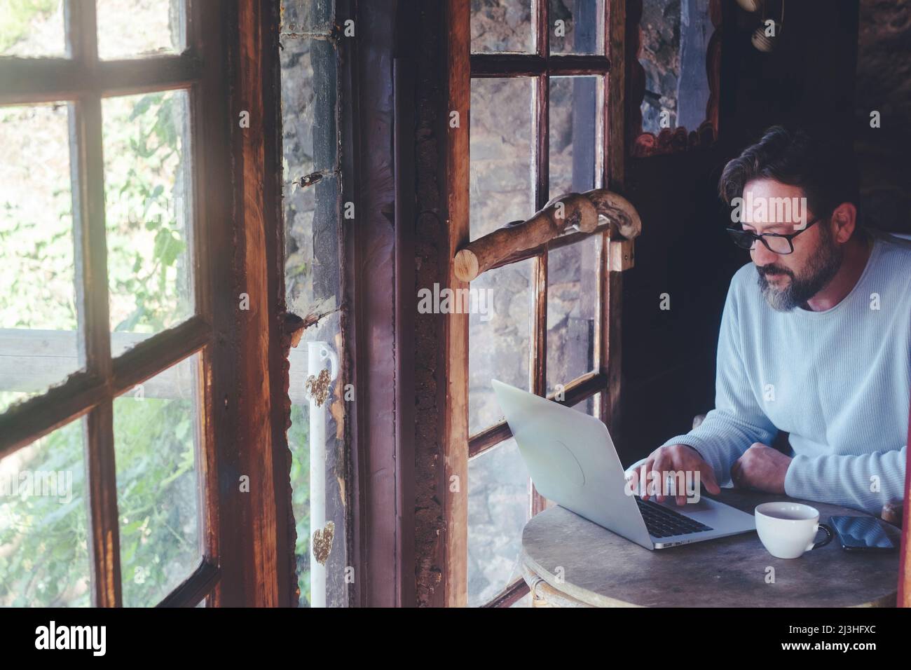 Mature man at work on laptop computer sitting at the table inside a home cozy cabin with wooden windows - digital smart working adult people concept lifestyle Stock Photo
