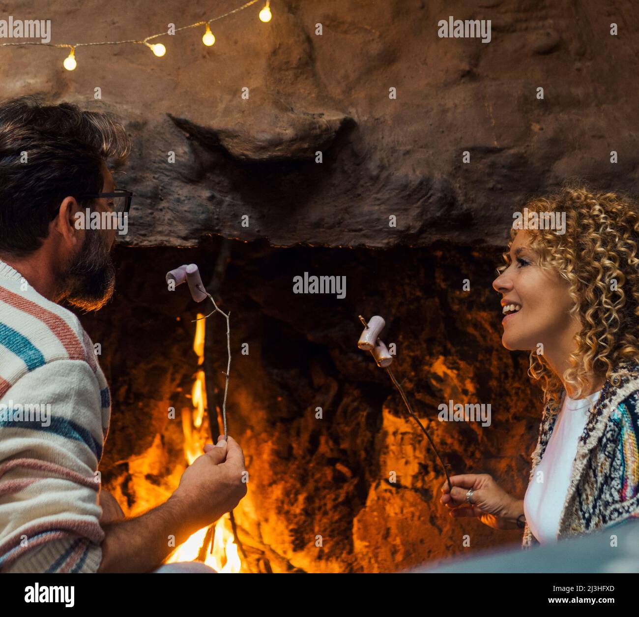 Happy couple eat marshmallows near a cozy fireplace flame at home in cozy home - festive celebration man and woman together with smile and happiness - adult people enjoying holiday vacation in winter season Stock Photo