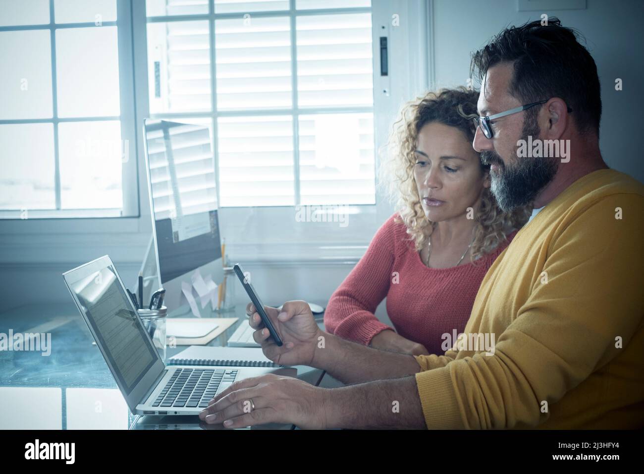 Smart working at home free office lifestyle with adult couple using home and two pc laptop computer on the desktop - modern man and woman online job remote workers bright day light Stock Photo