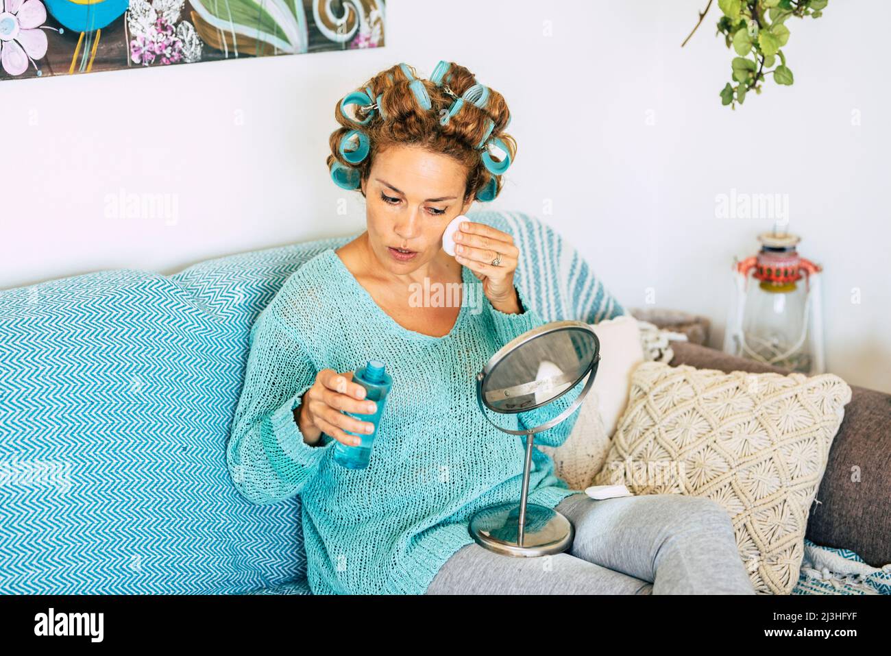 Attractive adult lady at home taking off make up and having care of her culery hair with curlers - blue colors - healthy care sking concept lifestyle with nice woman cleaning her face looking on a mirror Stock Photo