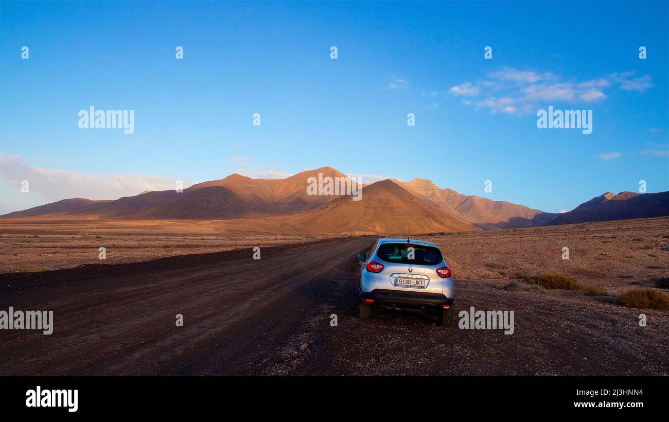 Spain, Canary Islands, Fuerteventura, southwest tip, barren landscape, silver car on dirt road, reddish brown lava hills in background, sky blue with few clouds Stock Photo