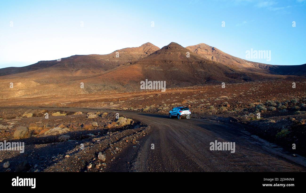 Spain, Canary Islands, Fuerteventura, southwest tip, barren landscape, dirt road, single silver car on dirt road, hills in background, sky blue Stock Photo