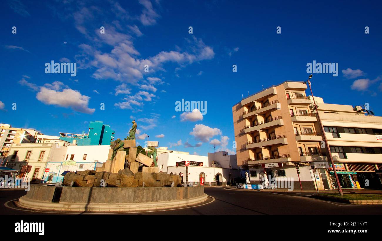 Spain, Canary Islands, Fuerteventura, capital, Puerto del Rosario, traffic circle island at the harbor promenade with sculpture, wide angle shot, skyscraper on the right, more buildings in the background, sky blue, clouds white Stock Photo