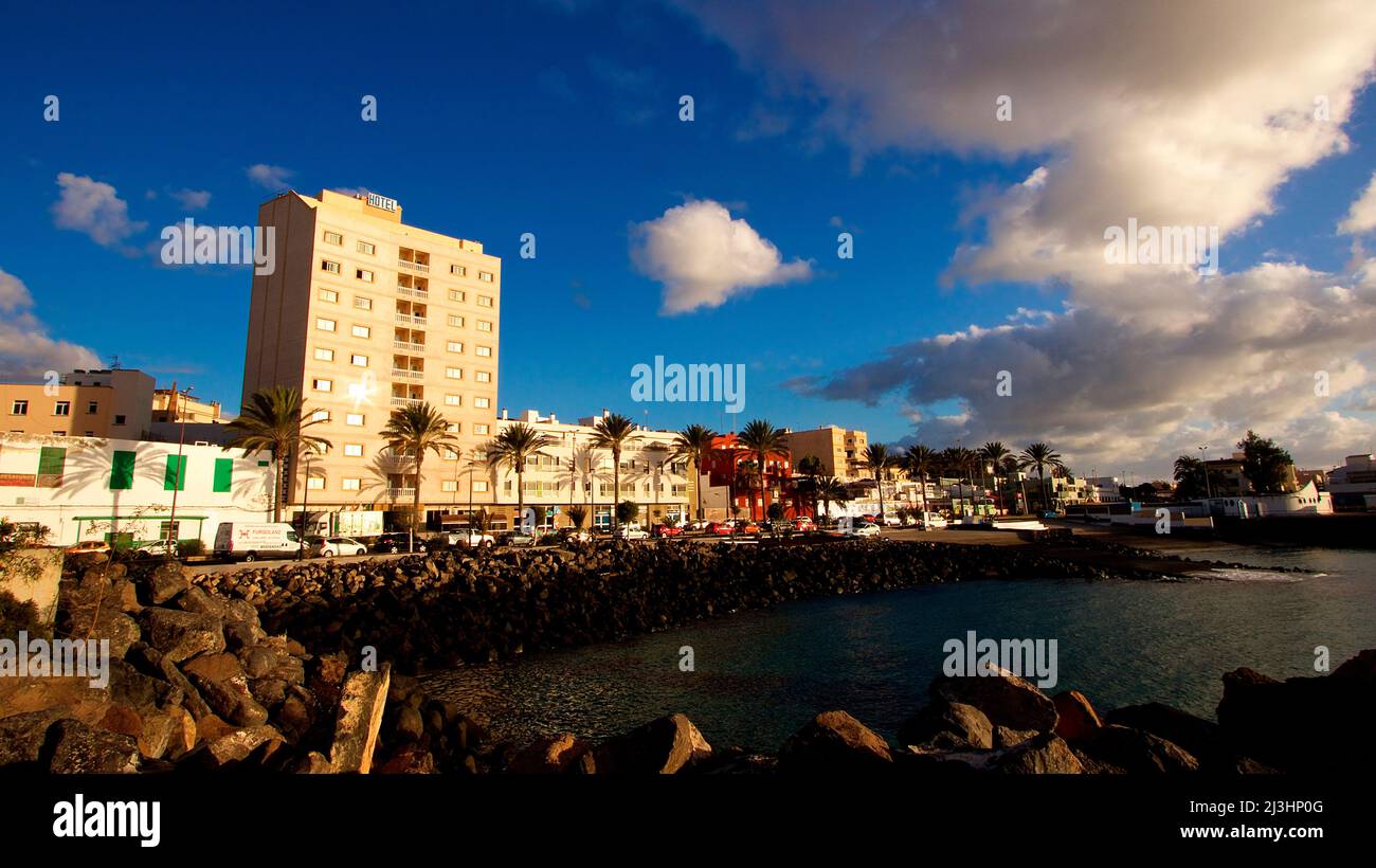 Spain, Canary Islands, Fuerteventura, capital, Puerto del Rosario, view over the water of the harbor to harbor promenade and row of buildings behind, a high-rise building, sky blue, clouds gray white Stock Photo