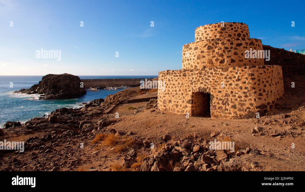 Spain, Canary Islands, Fuerteventura, west coast, Punta de Toston, three-tiered angular tower on the right of the picture, sea and rocks and surf on the left, blue sky, few white clouds Stock Photo