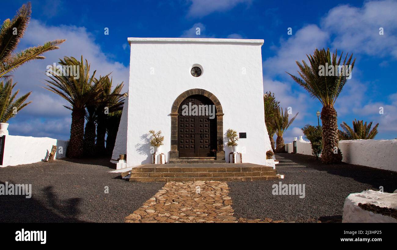 Canary Islands, Lanzarote, volcanic island, northwest coast, hermitage, church, Ermita de las Nieves, sky blue, white building of the Ermita, brown big door, stone framed, palm trees left and right, sky blue with white clouds Stock Photo