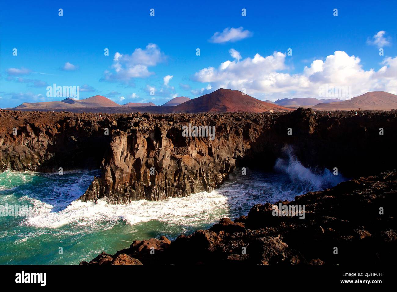 Canary Islands, Lanzarote, volcanic island, southwest coast, rugged volcanic coast, strong surf, sea caves, surf meets rugged coast, clear, blue sky, some white gray clouds, water frothing up, spray white Stock Photo