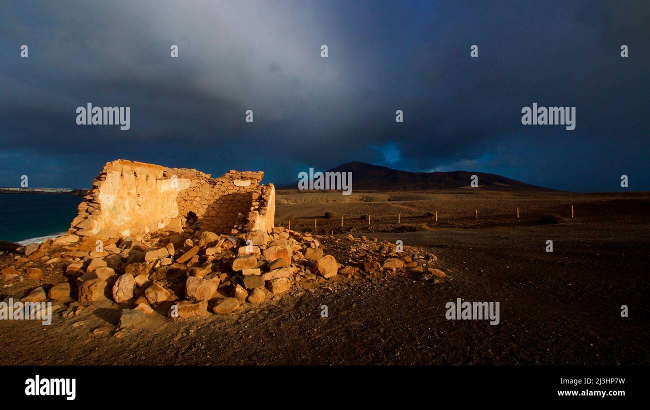 Canary Islands, Lanzarote, volcanic island, after-thunderstorm mood, Papagallo beaches, deserted, sky blue with clouds gray-white, morning light, ruins of an old building, walls, in the background black hills, sky dark and threatening, only the building is shone by the morning sun Stock Photo