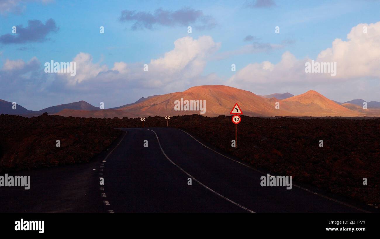 Canary Islands, Lanzarote, volcanic island, Timanfaya National Park, volcanic landscapes, an asphalt road in the shade leads to the national park, red-brown volcanic crater in the middle distance, blue sky, gray and white clouds, traffic sign '40', traffic sign 'Sharp left curve' Stock Photo