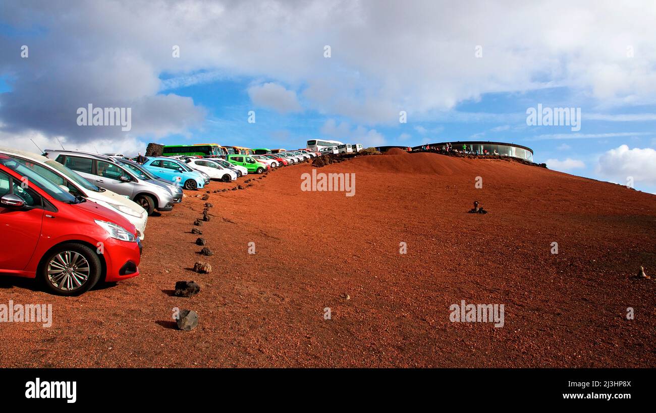 Canary Islands, Lanzarote, volcanic island, Timanfaya National Park, volcanic landscapes, Beuscherzentrum, round restaurant made of volcanic rock on the right in the background, blue sky above with white-gray clouds, in front of it cars parked in a long row on red lava earth Stock Photo