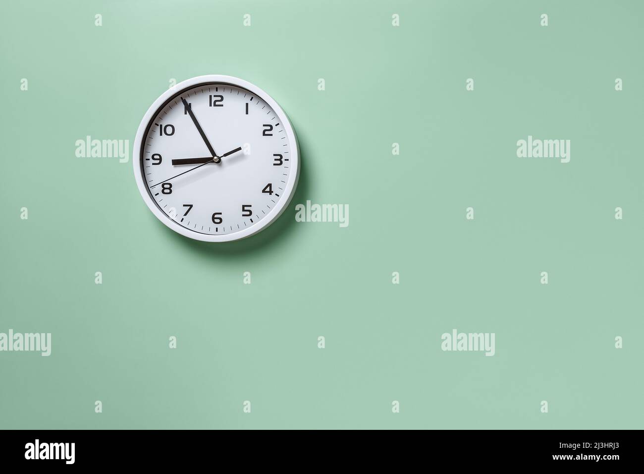 Wall clock hanging on the pale green wall with copy space. Round white clock with black hands. Five minutes to nine. Time measuring, hour and minutes. Stock Photo