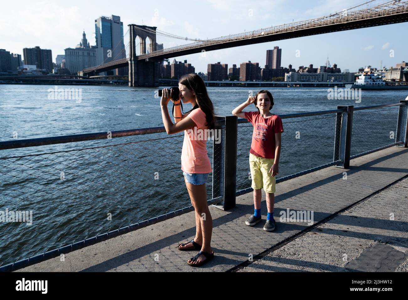 Dumbo/FULTON FERRY, New York City, NY, USA, 14 years old caucasian teenager girl and 12 years old caucasian teenager boy - both with brown hair and summer styling in front of the Brooklyn Bridge over East River Stock Photo
