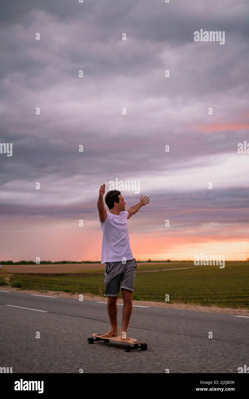 Freedom and active happy people concept lifestyle with young boy moving on skateboard lonboard on asphalt road outstretching arms agains a colorful am Stock Photo