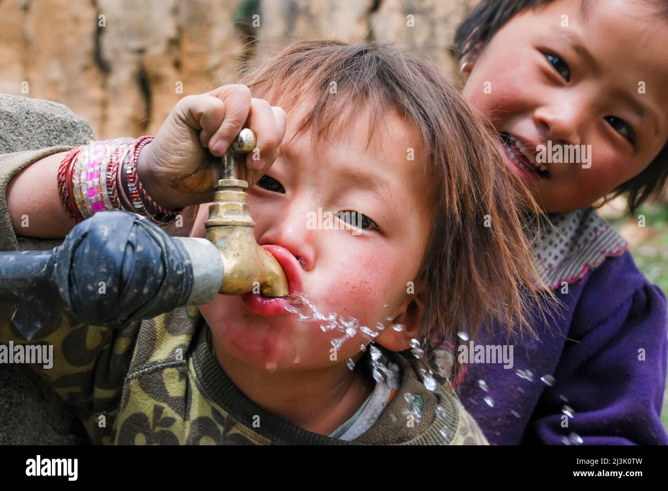 In a Phobjika village, a young girl drinks water from a spigot as another girl looks on; Phobjika, Bhutan Stock Photo