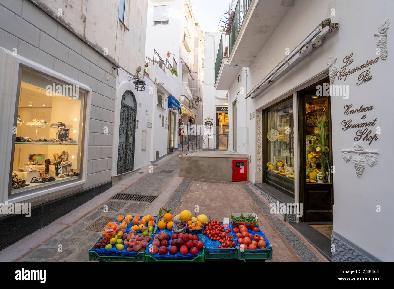 Fashion shops and a stall with fresh vegetables and fruit displayed in the narrow street in Capri; Capri, Naples, Italy Stock Photo