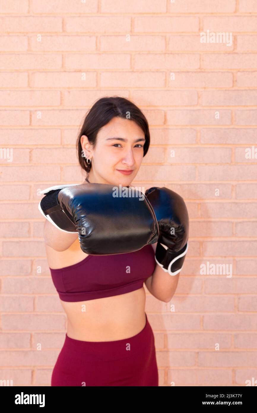 A young Caucasian woman wearing black boxing gloves is punching in front. The punching fist is in focus and the rest is out of focus. In the backgroun Stock Photo