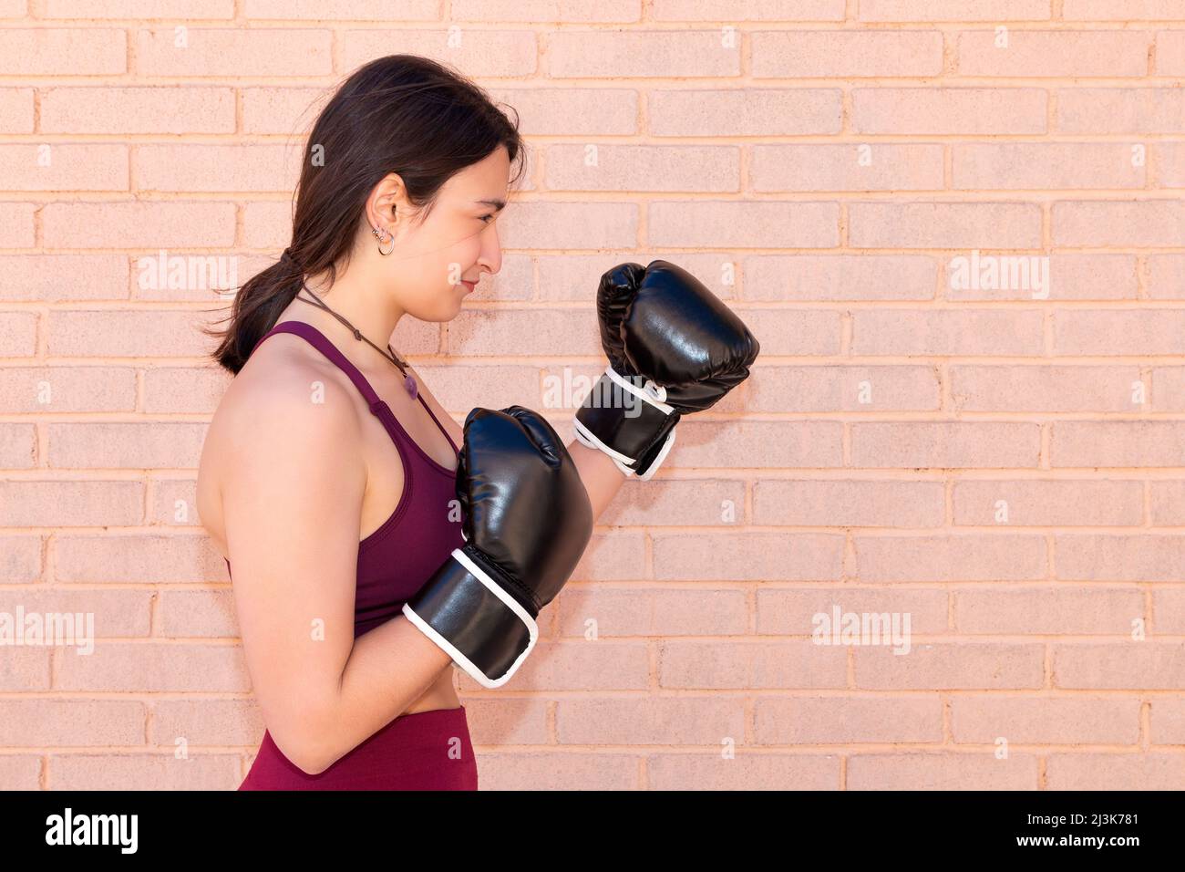 A young Caucasian woman wearing black boxing gloves in a defensive position seen from the side. In the background is a brick wall. Stock Photo