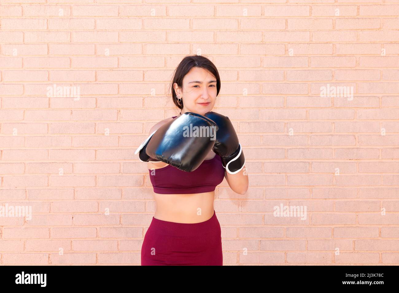 A young Caucasian woman wearing black boxing gloves is punching in front. The girl's face is in focus. In the background is a brick wall. Stock Photo