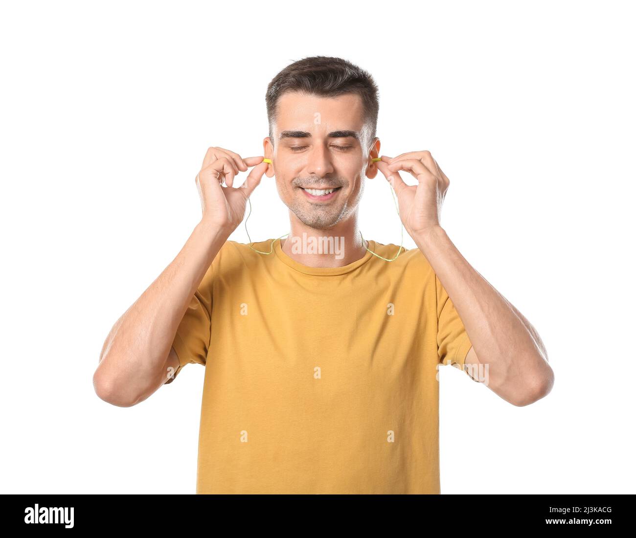 Smiling young man in yellow t-shirt putting earplugs on white background Stock Photo