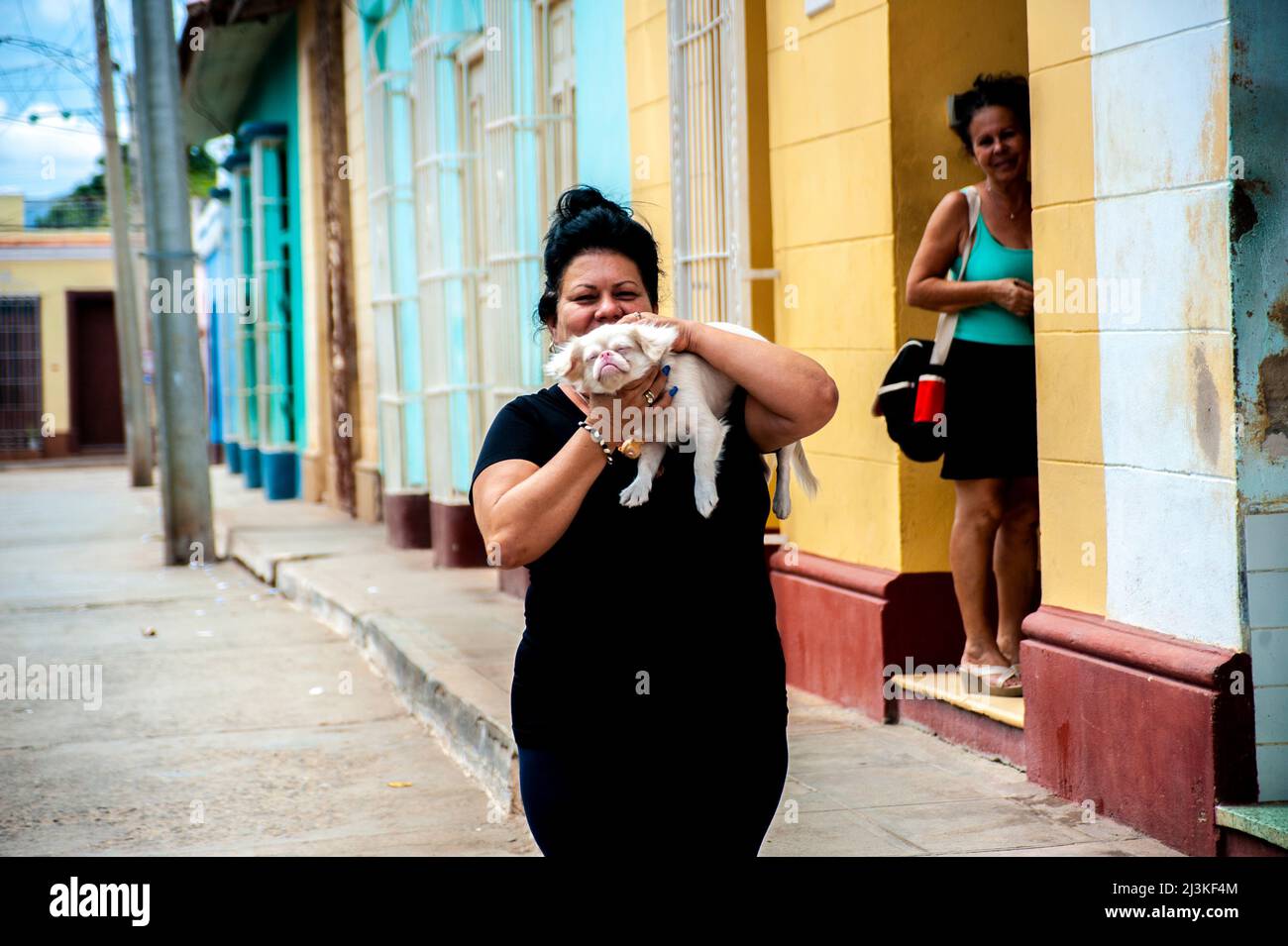 Woman holds and kisses her pet dog on a street in Trinidad, Cuba as her friend looks, smiles and laughs. Stock Photo