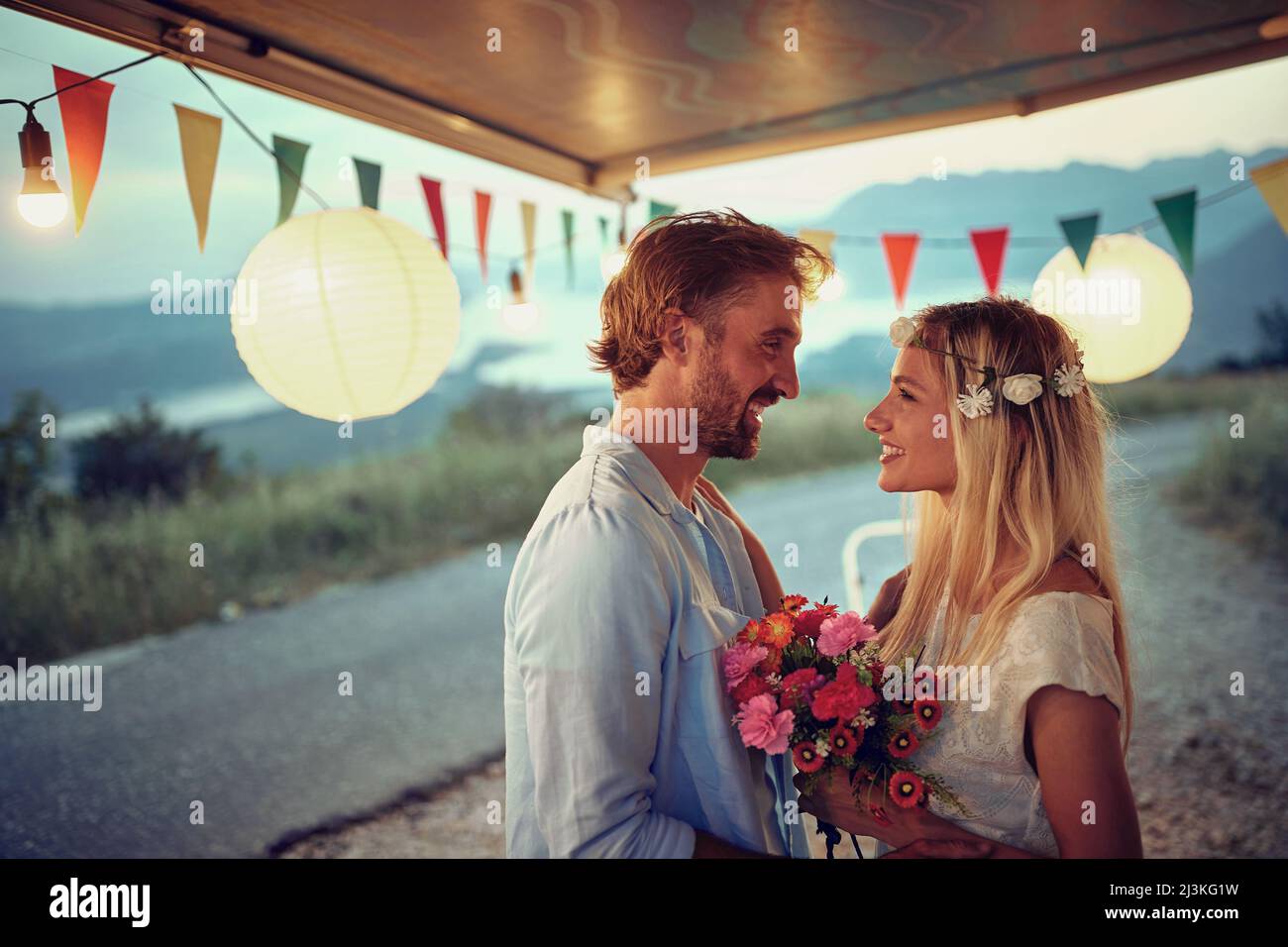 Boho style wedding couple at sunset in front of camper rv smilling at each other. Bride with bridal bouquet. Wedding ceremony, love, nature concept Stock Photo