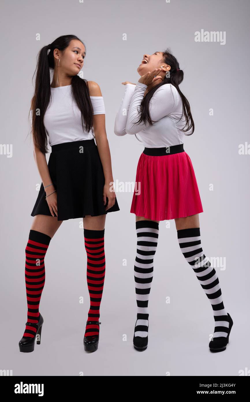 Two young and pretty girls, female models, wearing striped knee socks, black and red skirts and white blouses show playful emotions, posing against wh Stock Photo