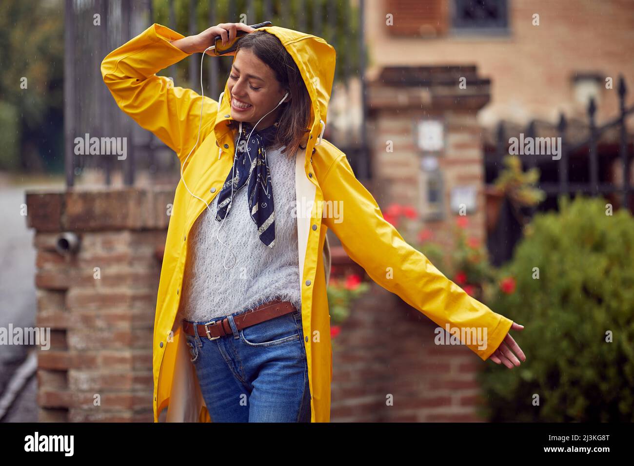 A young cheerful woman in a yellow raincoat is in ecstasy while listening to the music and walking the city in a good mood on a rainy day. Walk, rain, Stock Photo