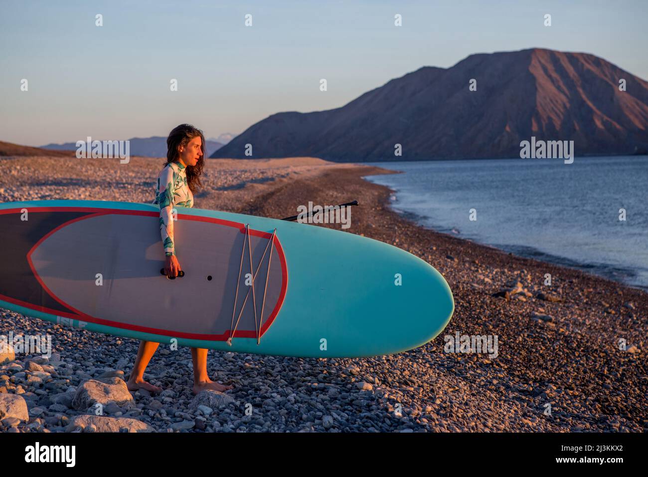 A woman carrying her paddleboard to the ocean on a remote beach on the Baja peninsula at sunrise. Stock Photo