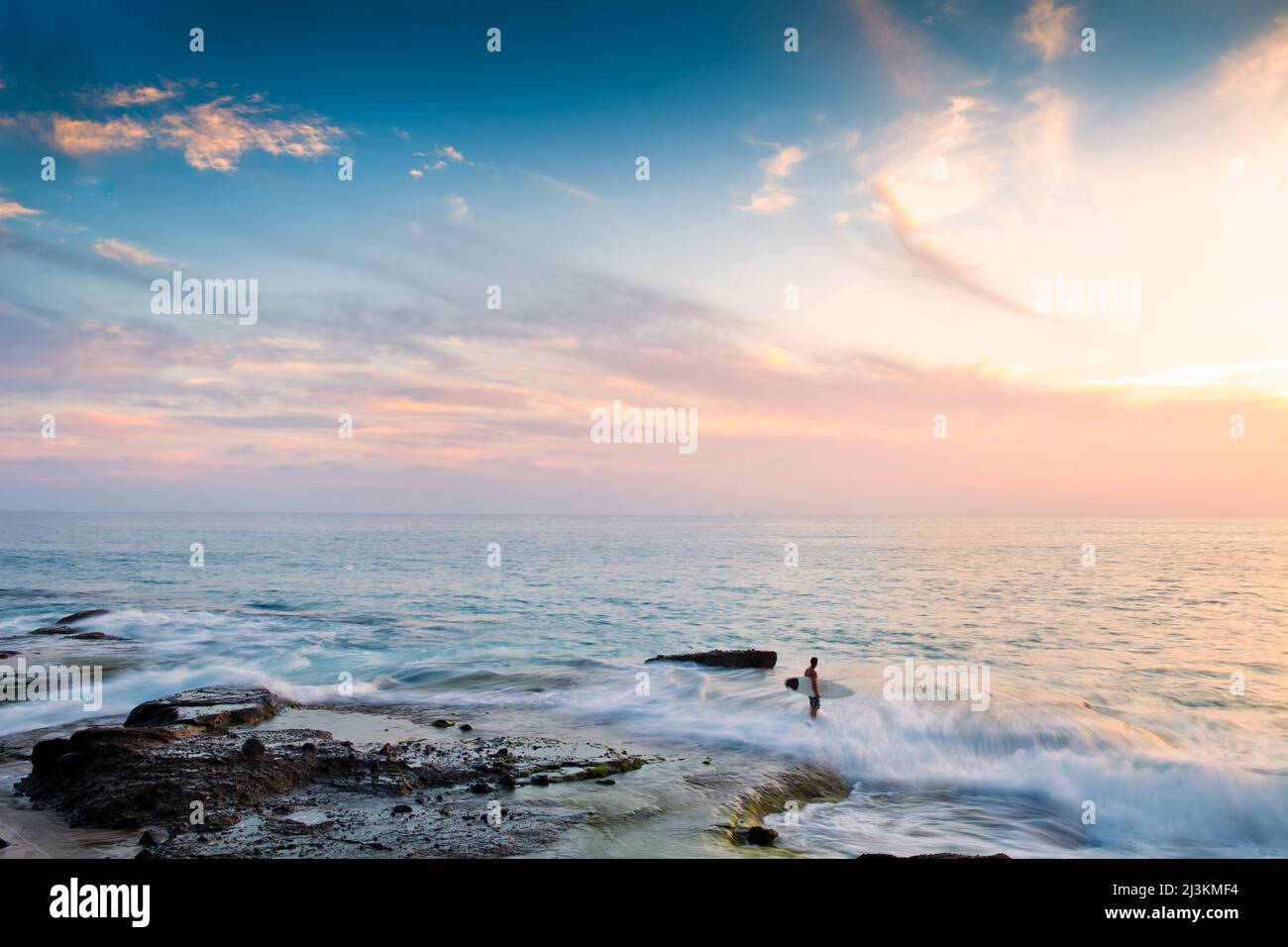 A surfer walking across the reef at sunset to access the waves. Stock Photo